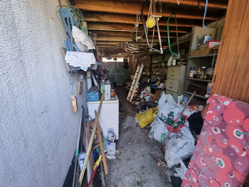 Long garage cluttered with shelving, tools, bags and timber before clearance