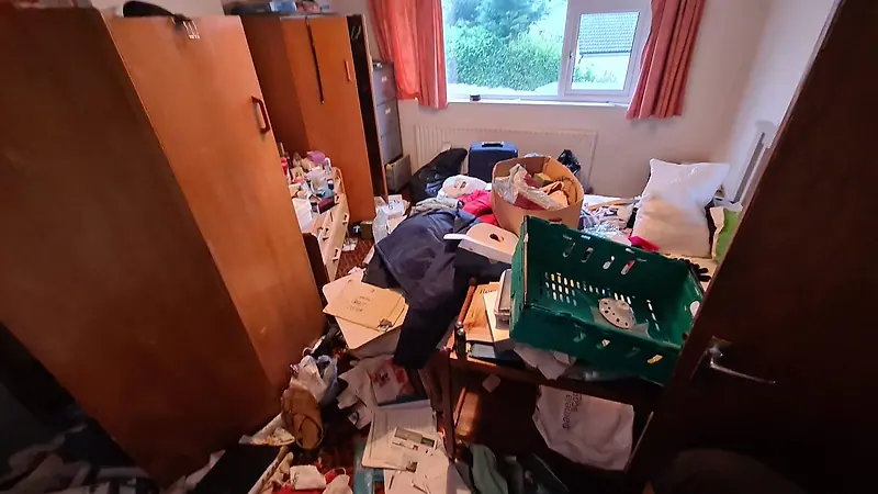 Front bedroom in Sutton before clearance, with clothes, crates and papers piled across the bed and floor